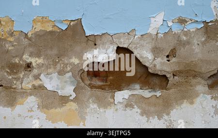 Horizontal close up of a decaying wall surface with peeling blue and white paint, cracked cement, exposed terra cotta colored layer, and red brick Stock Photo