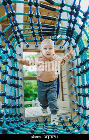 A child climbs through a rope tunnel in a playground. The child wears a brown hoodie and gray pants. The scene shows trees and a bright sky in the bac Stock Photo