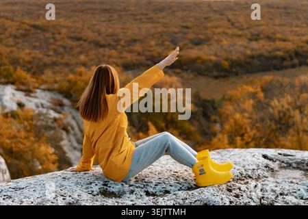 Woman autumn hiking sits on a rocky cliff watching the expansive forest valley view Stock Photo