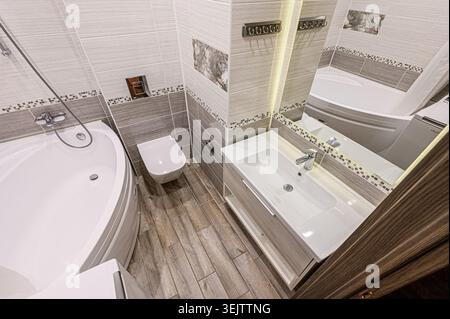 interior apartment Modern, compact bathroom with white bathtub, toilet, and shower.  Neutral grey and white tile, minimalist design. Clean lines and s Stock Photo