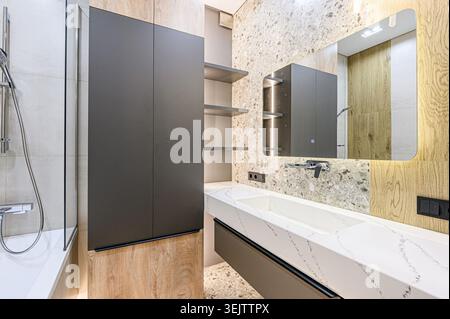 interior apartment Modern bathroom with wood and grey accents.  Features a sleek bathtub, glass shower enclosure, floating vanity with integrated sink Stock Photo