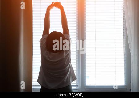 Woman standing with arms raised, stretching her body in front of a window with blinds, enjoying the warm light of sunrise awakening her mind and Stock Photo