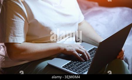 Woman hands working on a laptop computer at home during nighttime, typing on the keyboard while sitting on a bed, focusing on remote work or Stock Photo