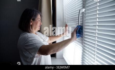 Woman carefully wiping dust off white horizontal window blinds using a blue microfiber cloth, performing a domestic chore to maintain a clean home Stock Photo