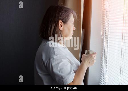 Mature woman standing indoors, looking through window blinds with sunlight streaming in, holding a glass of water and contemplating while finding Stock Photo