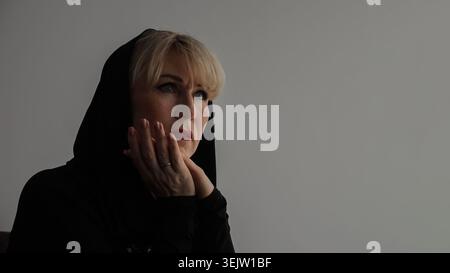 Grieving mature woman covering her face with hands and wearing a dark hooded garment, showing deep sorrow and emotional pain with a thoughtful Stock Photo