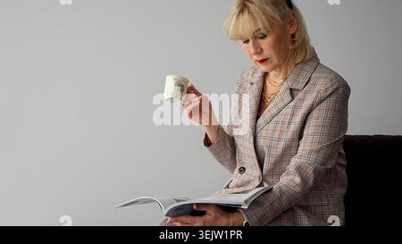 Mature executive woman in a plaid blazer relaxes indoors with a magazine and patterned coffee cup, stylish and confident during a thoughtful break Stock Photo
