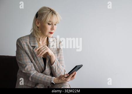 Senior blonde businesswoman wearing a plaid suit and golden necklace, sitting and concentrating on a smartphone, representing modern communication Stock Photo