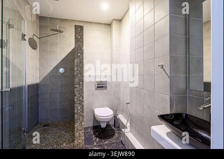 Modern, sleek bathroom with gray tile, glass shower enclosure, black sink, and compact toilet.  Marble-like flooring adds a touch of luxury. Minimalis Stock Photo