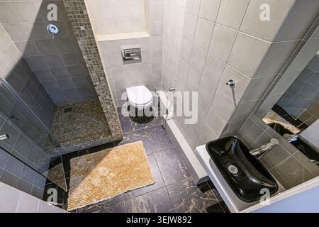 Modern, sleek bathroom with gray tile, glass shower enclosure, black sink, and compact toilet.  Marble-like flooring adds a touch of luxury. Minimalis Stock Photo