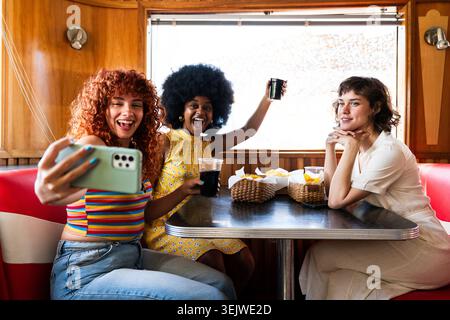 Multiethnic group of young happy friends bonding at vintage diner burger house restaurant - three happy young women enjoying food and drinks at a retr Stock Photo