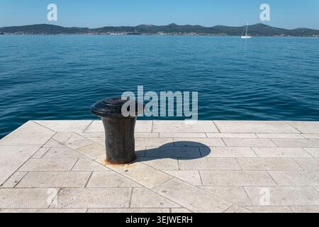 A solitary black metal mooring bollard on a stone pier, overlooking a serene blue sea with a distant sailboat and coastal hills. Stock Photo