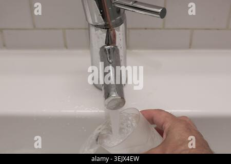Water flows steadily from chrome faucet. Clean white sink and tiled background. Simple, everyday action captured clearly. Ideal for home, kitchen Stock Photo