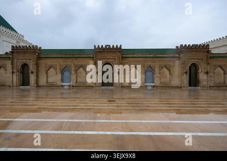Mausoleum courtyard in Rabat, Morocco, with ornate carved walls, green tiled rooflines, wet paving, and symmetrical Moorish architectural details. Stock Photo