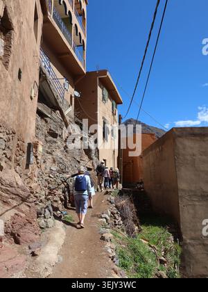 Tourists follow a local guide on a hiking trail through the village of Imlil in the foothills of the Atlas Mountains and pass local homes & buildings. Stock Photo