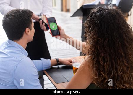 Rear view of a couple paying a restaurant bill with a smartphone using contactless technology. Waiter holds the payment terminal while the customer co Stock Photo