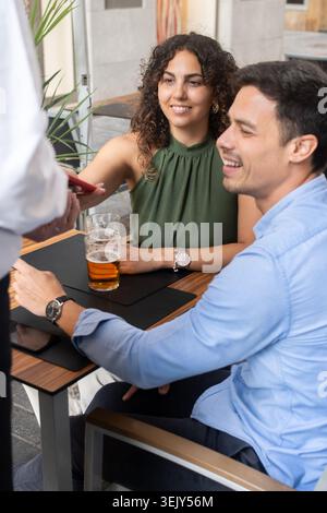 Happy young couple sitting at an outdoor restaurant terrace while making a contactless mobile payment. Waiter holds the payment terminal as the woman Stock Photo