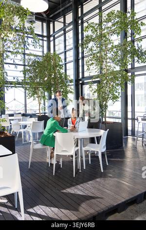 Diverse coworkers collaborating around round white table in glass atrium using laptop, phones Stock Photo