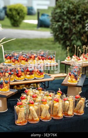 The photograph showcases beautifully arranged portioned snacks in small glasses and fresh fruit cups on wooden stands during an outdoor event Stock Photo