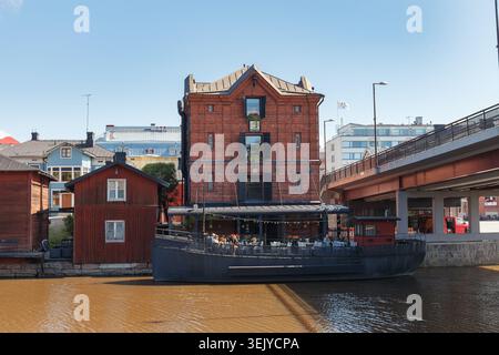 PORVOO, FINLAND - AUGUST 15, 2025: Red brick warehouse building with restaurant terrace beside river bridge in nordic town under bright summer sky. Stock Photo
