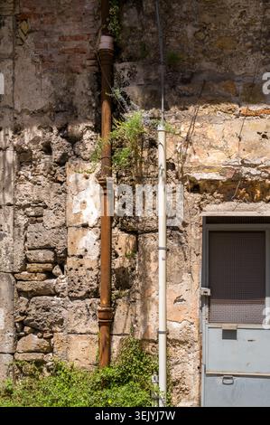 This weathered, historic stone wall displays urban decay as old, rusted pipes and modern conduits run alongside a gray door, highlighting the passage Stock Photo