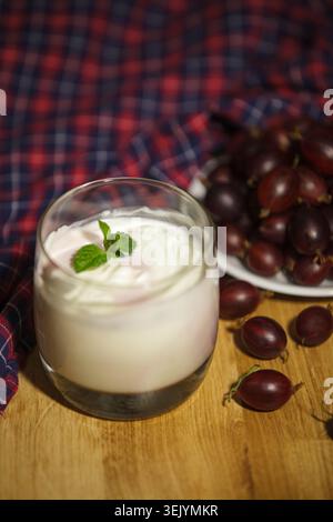 Vertical shot of a healthy natural yogurt decorated with mint in a glass. Ripe red gooseberries on a plate and checkered cloth in the blurred backgrou Stock Photo