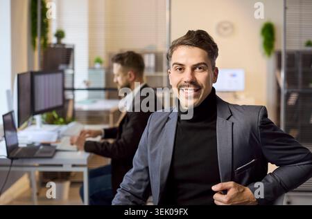 Smiling confident businessman portrait in modern office with coworker at computer Stock Photo