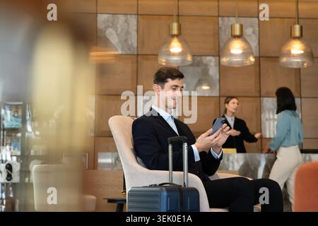 Male guest sitting in lobby with suitcase, using smartphone Stock Photo