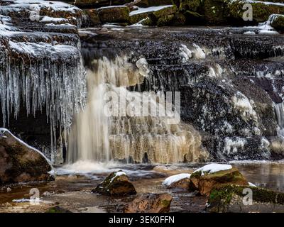 View of heavy icicles hanging from rocky banks above a frozen river Stock Photo