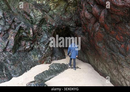 A woman entering a natural cave made of Cornish Serpentine - a rare, richly hued red & green rock found only on the Lizard Peninsula in Cornwall, UK Stock Photo