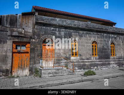 Old stone building with weathered wooden doors and windows. Blue sky above, empty street below. Structure shows signs of decay and abandonment Stock Photo