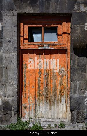 Old orange door with peeling paint, set in stone wall. Small window above door frame. Moss grows at base of door. Sunlight highlights weathered wood t Stock Photo