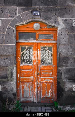 Weathered orange wooden door with peeling paint. Stone wall surrounds arched doorway. Door number 109 above. Fan-shaped glass panes visible. Green mos Stock Photo