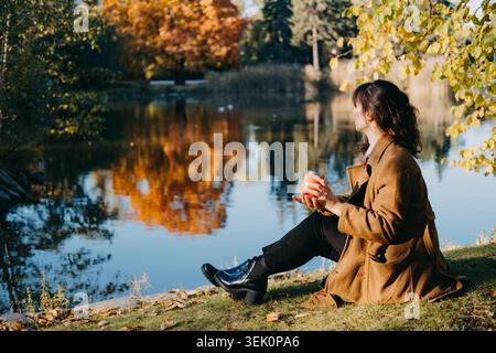 Woman sits holding cup on grassy bank by pond in autumn park. Mindful recreation by water, outdoor wellness lakeside destination, mental wellbeing Stock Photo