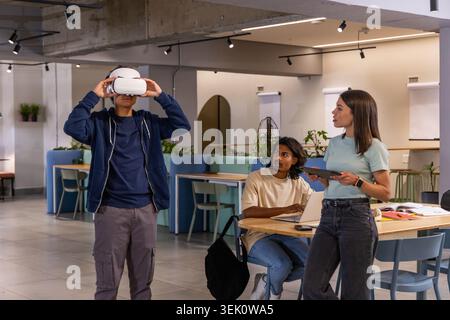 Diverse coworkers testing virtual reality headset at co-working table with laptop and tablet Stock Photo