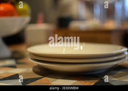 Close-up of three stacked white ceramic plates on a decorative surface with blurred showing colorful fruit and kitchen items. Stock Photo
