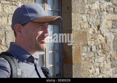 A man wearing a gray cap and button-up shirt with a backpack stands next to a stone wall and window. The wall is composed of rough, light-colored ston Stock Photo