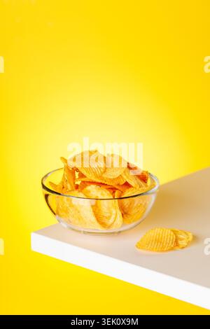 Glass bowl with ridged potato chips on white pedestal against bright yellow background, minimal snack concept Stock Photo