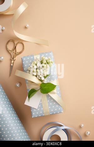 Gift box in light blue polka dot paper with cream satin ribbon, white lilac flowers, blank tag, gold scissors and pearls on beige background. Copyspac Stock Photo