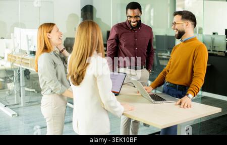 Four team members engage in conversation and share smiles in a modern office. They stand around a table with laptops open, discussing work and enjoyin Stock Photo