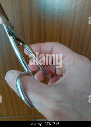 Side view of a Caucasian man's hand holding and using classic silver metallic scissors against a blurred wooden background. Stock Photo