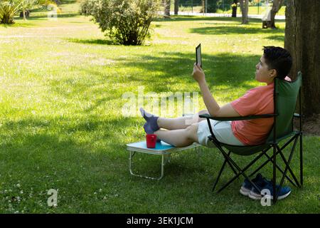 A young person relaxing outdoors with an e-reader in a park, with their legs propped up on a table Stock Photo