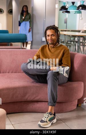 African American coworkers sitting in lounge on pink sofa, man holding tablet woman using phone Stock Photo