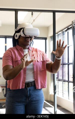 African American woman wearing white VR headset and pink shirt using watch operating VR in office Stock Photo