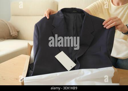 Woman holding new dark jacket taken out of a shipping box. Online clothes shopping concept in home interior. Middle aged woman unboxing package with Stock Photo