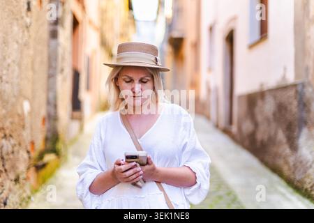 Middle aged woman using smartphone while walking through narrow historic European street Stock Photo