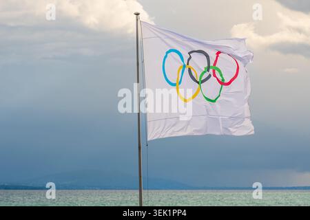 White Olympic flag waving on a flagpole above Lake Geneva with dramatic cloudy sky, water and distant mountains in Lausanne, Switzerland. Stock Photo
