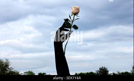 A black arm monument reaches upward toward an overcast sky holding a delicate light pink hybrid tea rose Rosa x hybrida blossom in its fingers Stock Photo