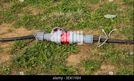 A close-up shot shows a red and grey waterproof cee industrial power cable connector link lying on an outdoor grassy patch with attached safety string Stock Photo