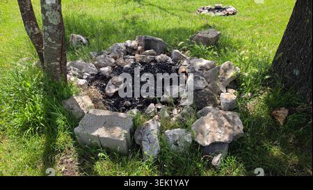 A rustic campfire pit made from irregular natural rocks and a concrete block sits surrounded by green grass and trees with leftover black charcoal ash Stock Photo